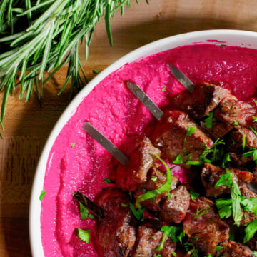 Grilled beef skewers served on a vibrant beet purée, garnished with fresh herbs and presented on a white plate with rosemary in the background.